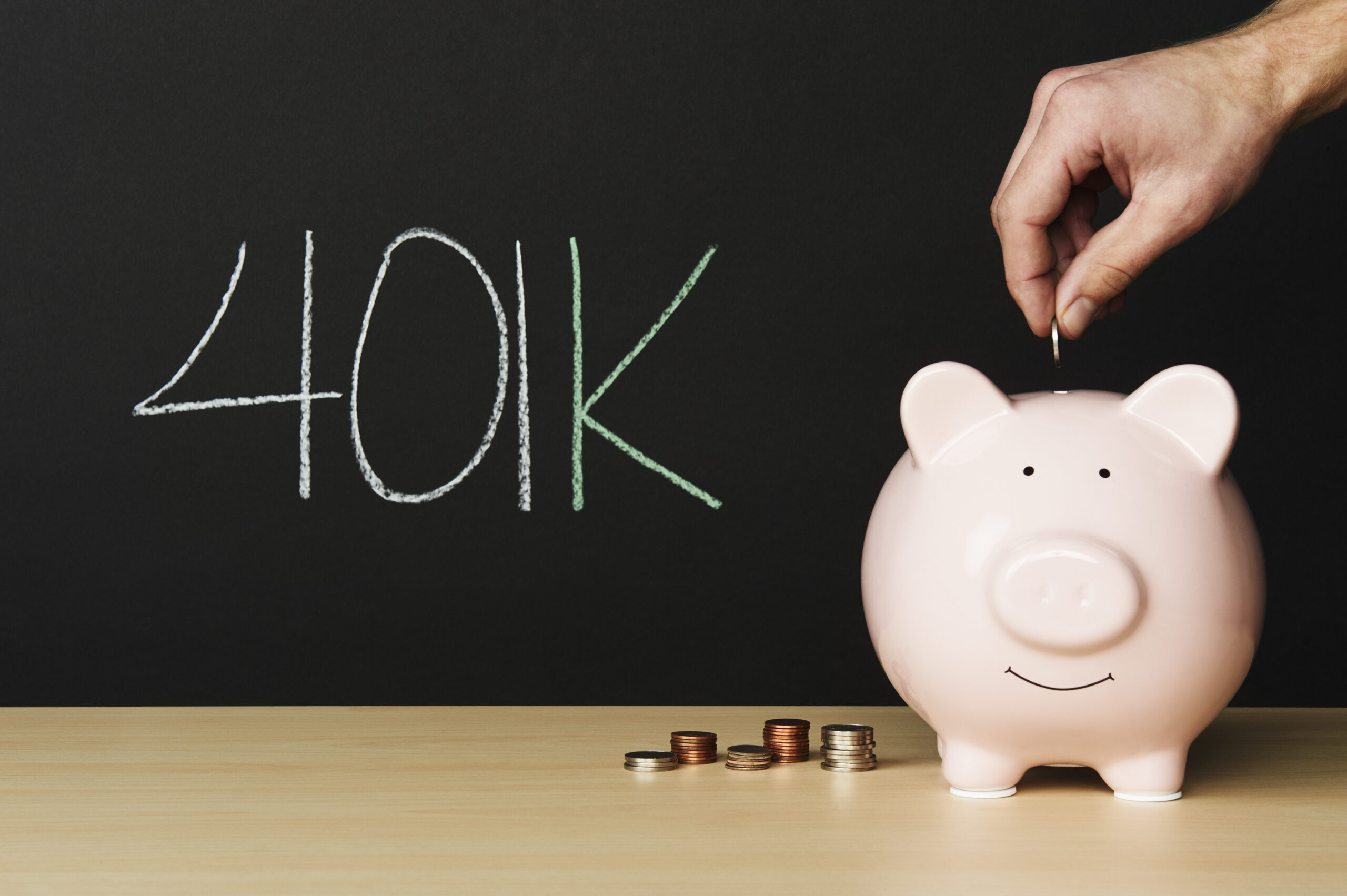 Male hand placing a coin into a piggy bank with 401k scrawled on a chalkboard in the background.